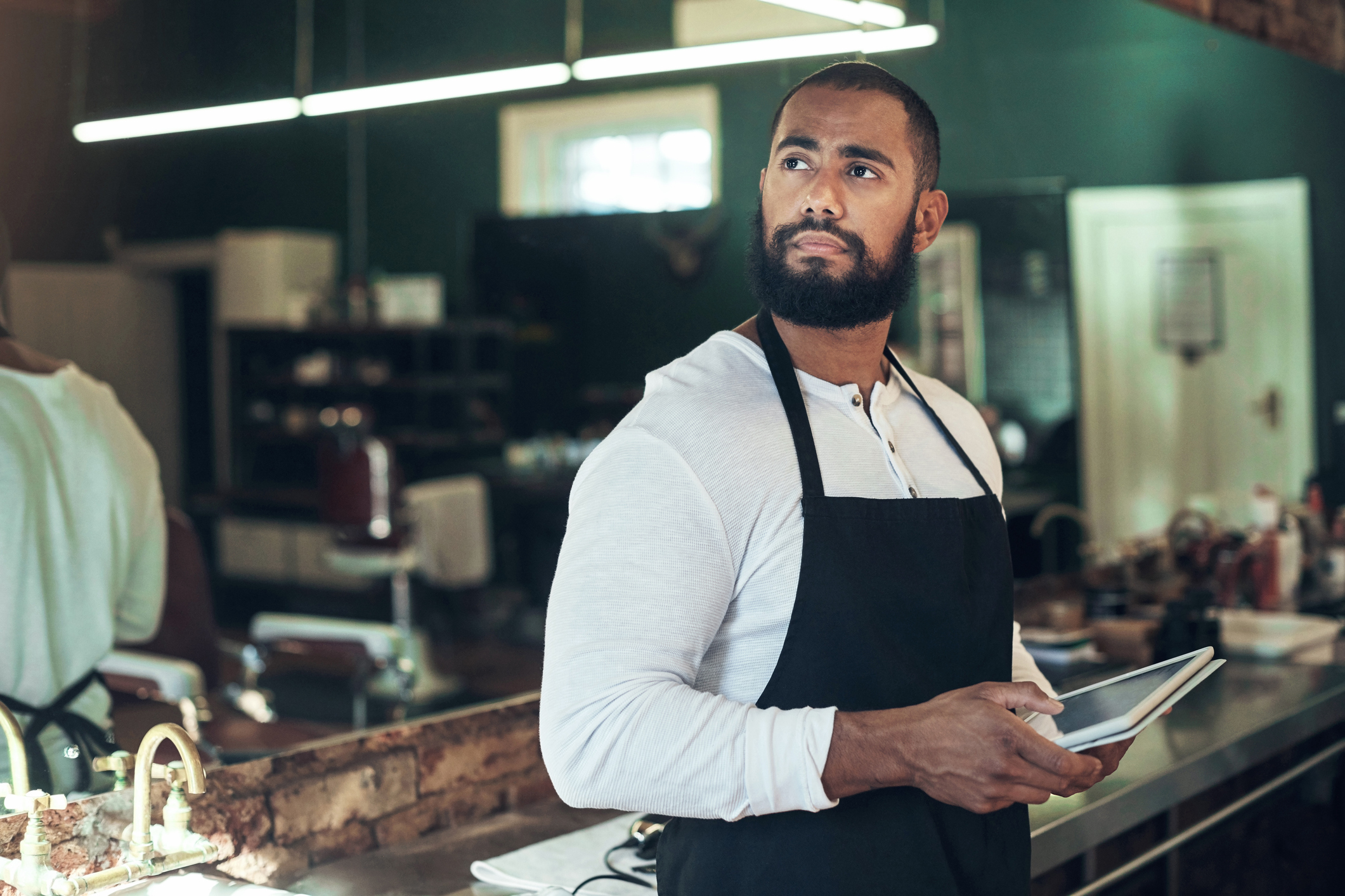 Shot of a handsome young barber standing alone and looking contemplative while using a digital tablet in his salon